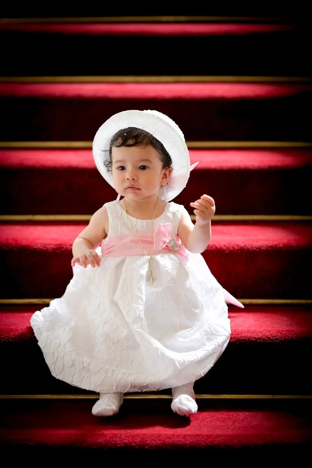 a baby in a white dress and hat on red carpet