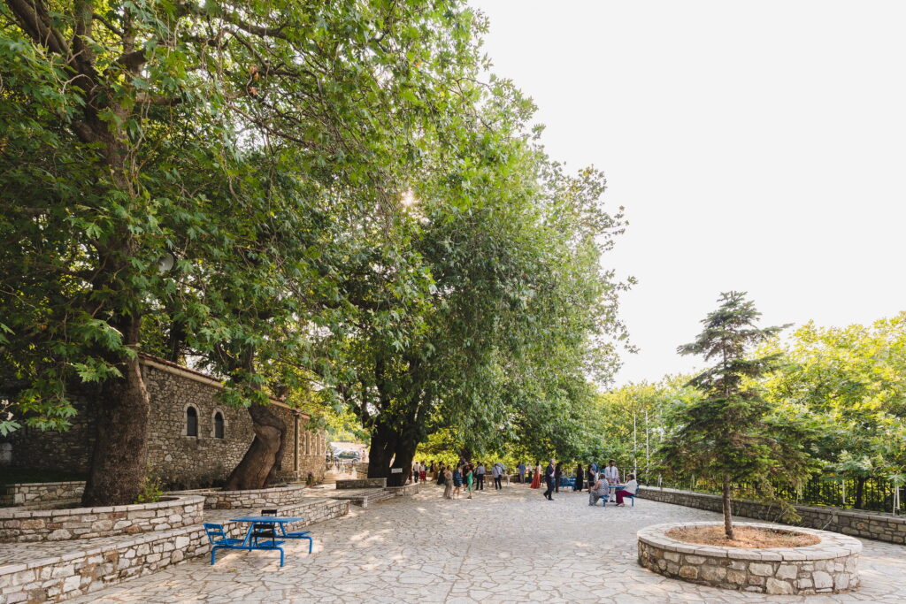Άνθρωποι συγκεντρωμένοι σε πλακόστρωτη αυλή με μεγάλα πλατάνια δίπλα σε πέτρινο κτίριο