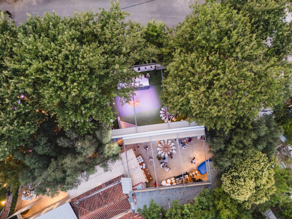 Άποψη από ψηλά ενός υπαίθριου χώρου εκδηλώσεων με δέντρα, τραπέζια και μια μωβ πίστα χορού